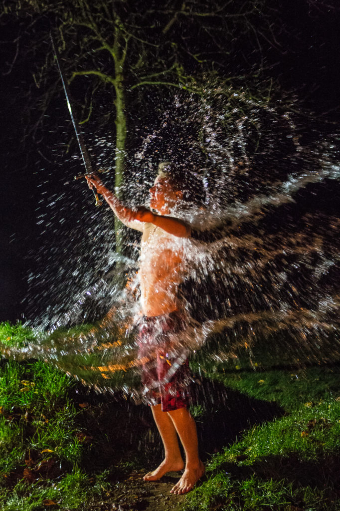 Ein Junge steht in Badehose bei Nacht auf einer Wiese und wird mit Wasser übergossen.